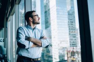 Contemplative male entrepreneur with crossed hands standing near office window view and feeling pondering during work day in company, Caucasian pensive corporate boss thoughtful looking away